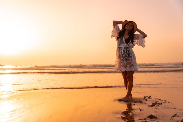 A woman in the sunset in a white dress at the sea in summer with a hat