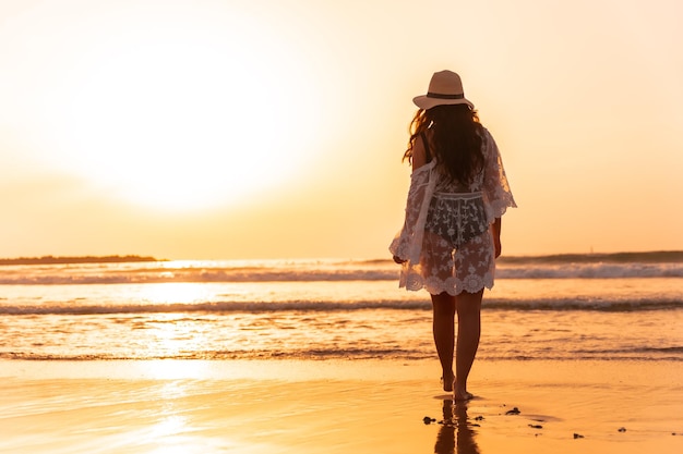干潮時に海を歩いている帽子をかぶった白いドレスを着て日没の女性