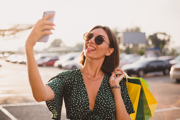 Woman at sunset with colorful shopping bags and parking lot by shopping mall happy with mobile phone take photo selfie