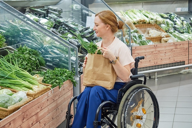 Woman in a supermarket chooses vegetables herbs uses an ecofriendly bag sitting in a wheelchair