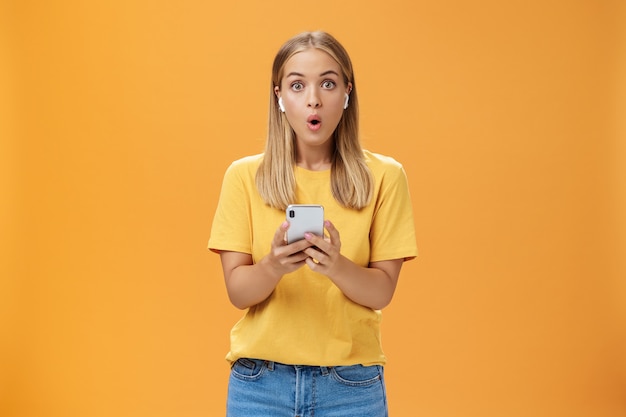 Woman surprised holding smartphone wearing earphones feeling impressed over orange background