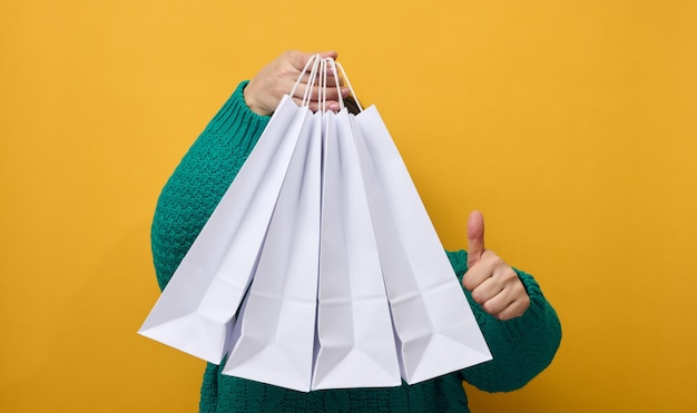 A woman in a sweater holds white paper bags on a yellow\
background and shows a like gesture with her hand. purchases.\
seasonal sale