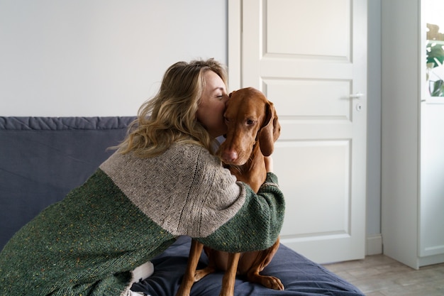 Woman in sweater hugging her beloved Wirehaired Vizsla dog, sitting on couch at home. Love pets.