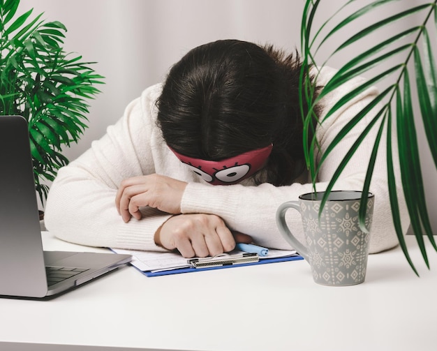 Woman in a sweater sleeps at a work table, next to a laptop. Fatigue and overwork concept. Laziness