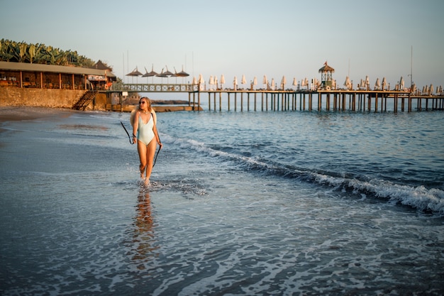 A woman in a swimsuit and a hat with glasses walks along the beach at sunset. The concept of sea recreation. Selective focus