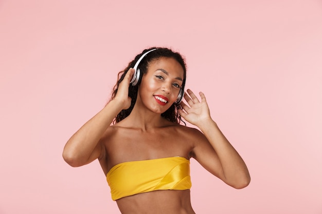 Woman in swimwear posing isolated over pink wall listening music with headphones