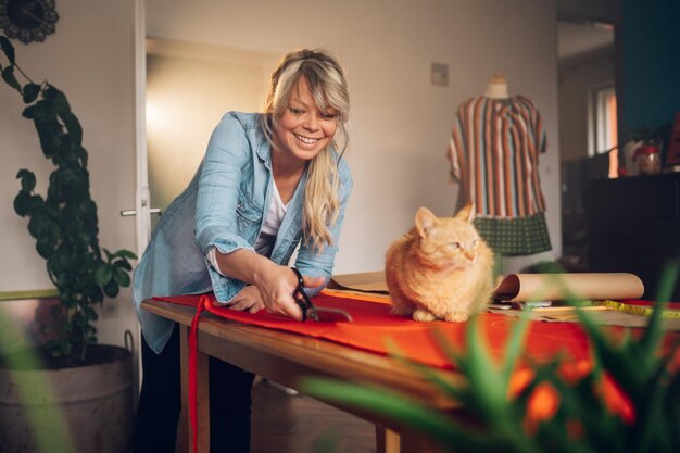 Woman tailor working from home and tailoring beautiful clothes\
with a pet cat