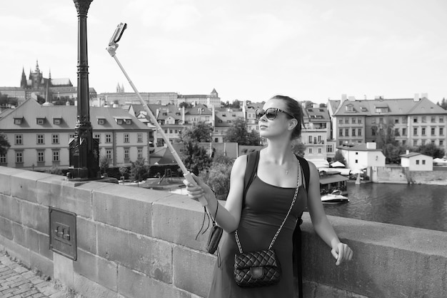 Woman take selfie with mobile phone in prague czech republic Woman with smartphone on stick at bridge over river Girl tourist with fashion look and sensual beauty Travelling and city sightseeing