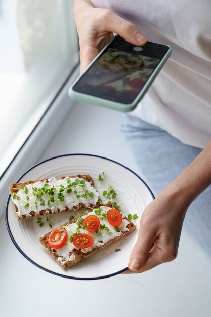 Woman takes a photo on smart phone, holding plate with rye crisp bread with cheese and micro greens