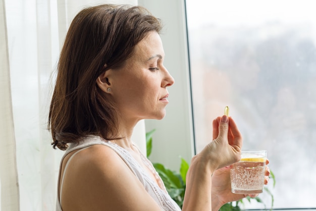 Woman takes pill with omega-3 and holding a glass of fresh water with lemon.