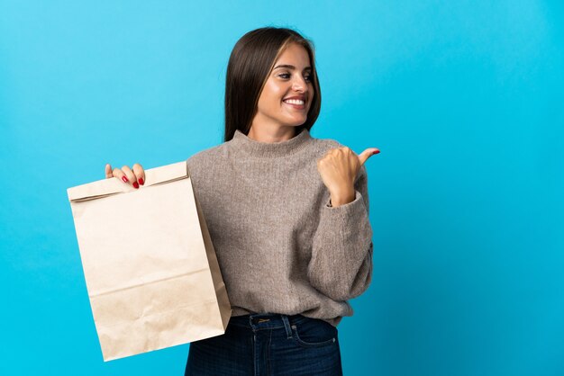 Woman taking a bag of takeaway food on blue pointing to the side to present a product