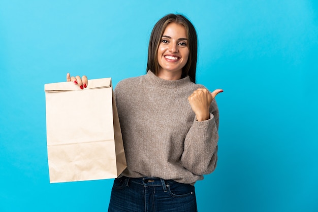 Woman taking a bag of takeaway food isolated on blue pointing to the side to present a product