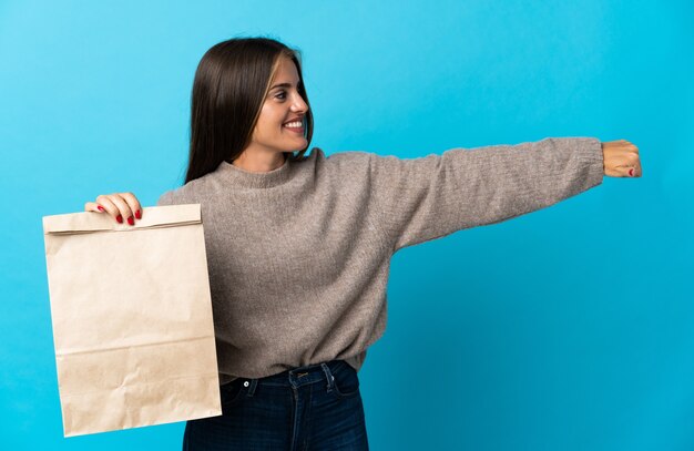 親指を立てるジェスチャーを与える青い壁に隔離された持ち帰り用食品の袋を取っている女性