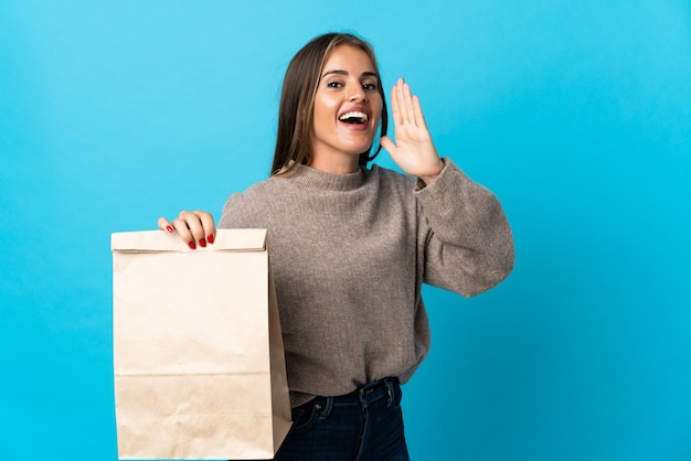 Woman taking a bag of takeaway food isolated on blue wall shouting with mouth wide open