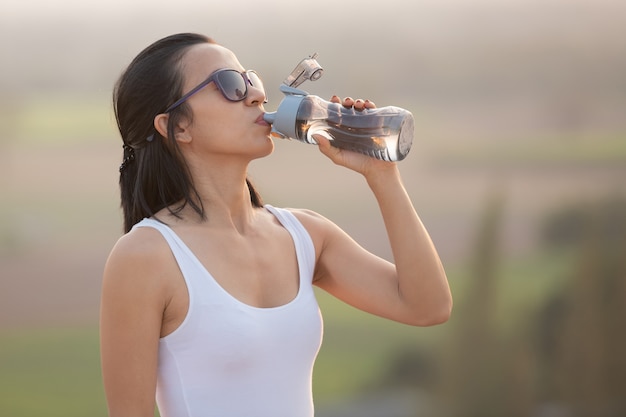 Woman taking a break to drink from water bottle while hiking and poles standing on a rocky mountain ridge looking out valleys.