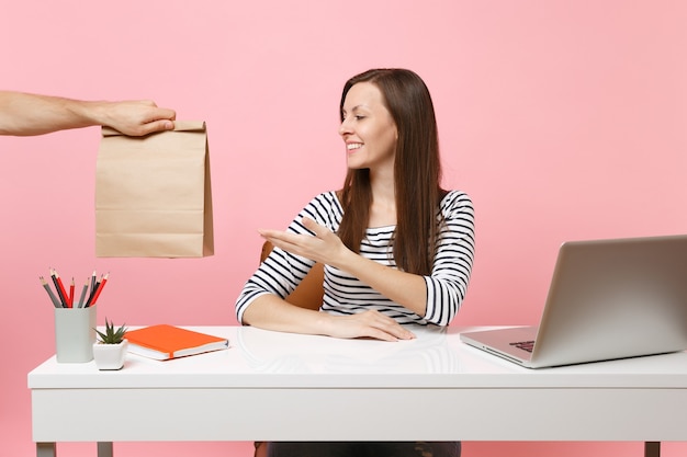 Woman taking brown clear empty blank craft paper bag, work at office with pc laptop isolated on pink background. Food products delivery courier service from shop or restaurant to office. Copy space.