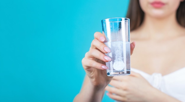 Donna che assume droghe per liberare il mal di testa bruna prende alcune pillole tiene un bicchiere d'acqua isolato sul blu