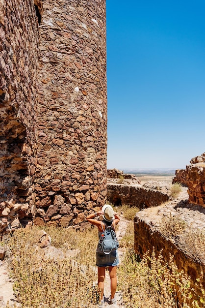 Woman taking his hat and standing in front of the defense stone\
walls of a medieval castle. almonacid castle, toledo