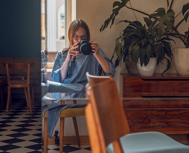 Женщина фотографирует себя, отражаясь в зеркале в современном кафе с дневным светом и зелеными растениями.