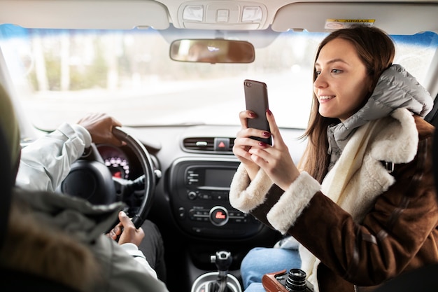 Woman taking picture of man with smartphone in the car while on a winter road trip