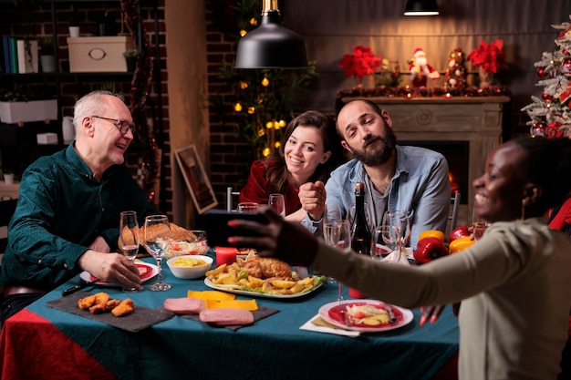 Woman taking portrait photo of diverse people at christmas festive dinner table. Big family gathering, wife and husband couple celebrating xmas with parents, drinking sparkling wine