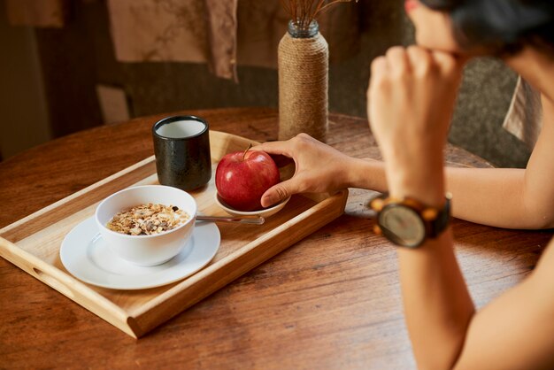 Woman taking red apple from tray with healthy breakfast consisting of porridge and cup of warm water