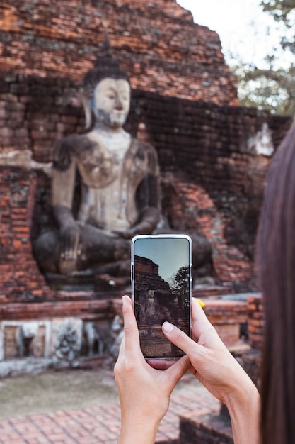 Foto donna che prende un'immagine verticale con il telefono dello schermo a buddha stante in wat mahathat nel parco nazionale di sukhothai.