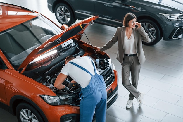 Woman talking by phone Man repairing automobile indoors Professional service