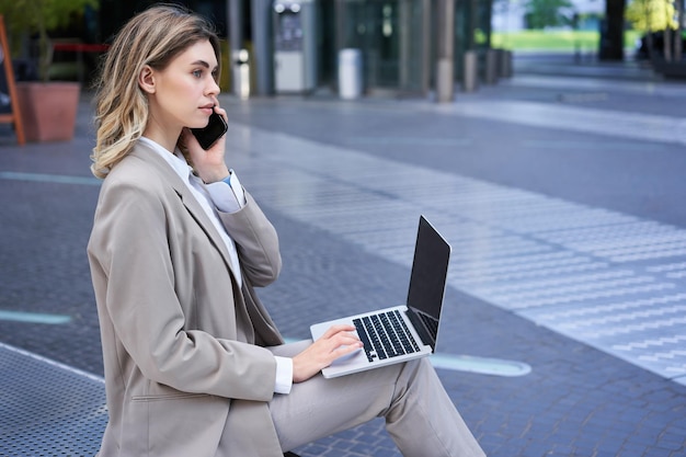 Woman talking on mobile phone and working with laptop sitting\
in city centre on street wearing suit