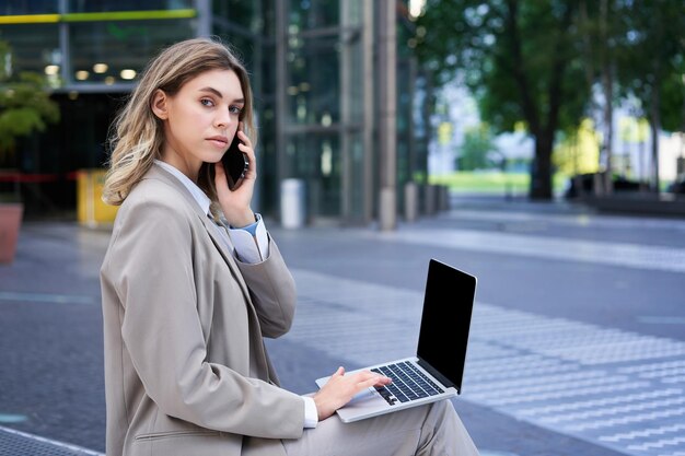 Woman talking on mobile phone and working with laptop sitting\
in city centre on street wearing suit