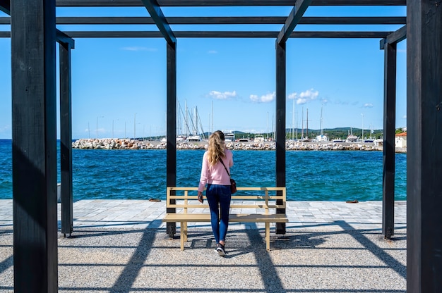 A woman talking on the phone in a gazebo on a pier with black metal posts and a bench, Aegean sea in Nikiti, Greece