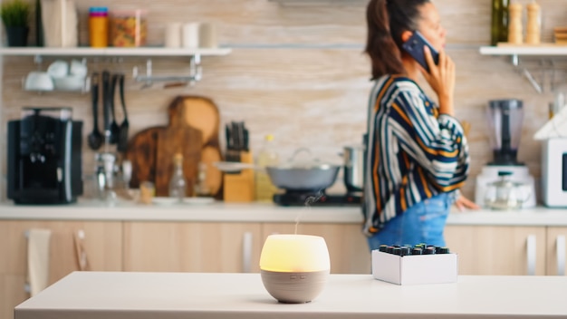 Woman talking on the phone with essential oils aromatherapy next to her in the kitchen. Aroma health essence, welness aromatherapy home spa fragrance tranquil theraphy, therapeutic steam, mental healt