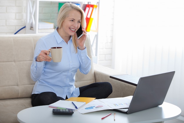 Woman talking on smart phone and smiling at her working place siting in the office.