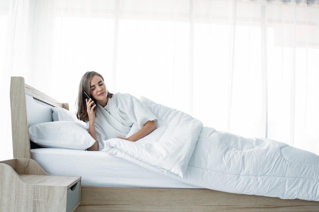 Woman talking smartphone on bed with a smiling face after waking up.