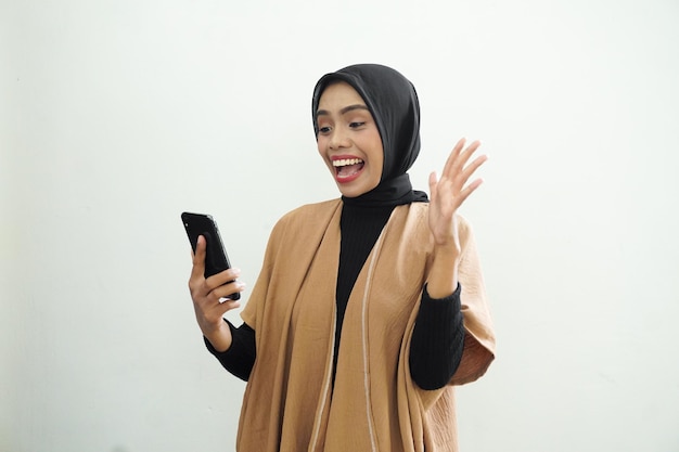 A woman in a tan jacket is holding a black phone and is laughing.