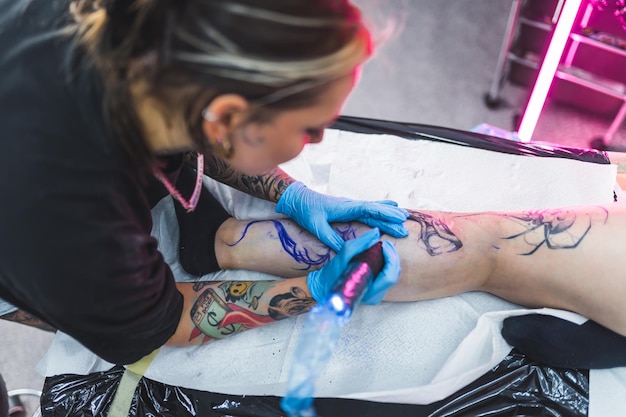 Woman tattoo artist drawing a tattoo on a clients leg concept art high angle indoor shot blurred