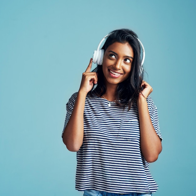 Думающая женщина и наушники с музыкой в студии со счастьем и веб-аудио Синий фон Индийская женщина и молодая модель слушают, слушают и транслируют песню с улыбкой и макетом