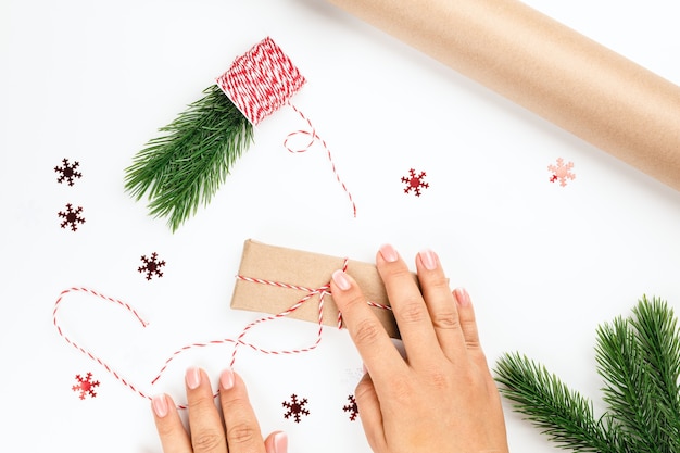 여자는 크리스마스 나무 가지와 빨간 눈송이로 장식된 선물 상자에 빨간색과 흰색 밧줄을 묶습니다.