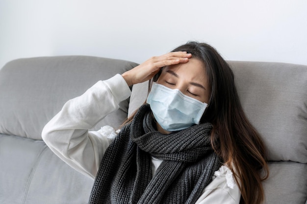 Женщина, касающаяся лба, с головной болью, лихорадкой, больная девушка в медицинской маске, сидящая дома на диване.