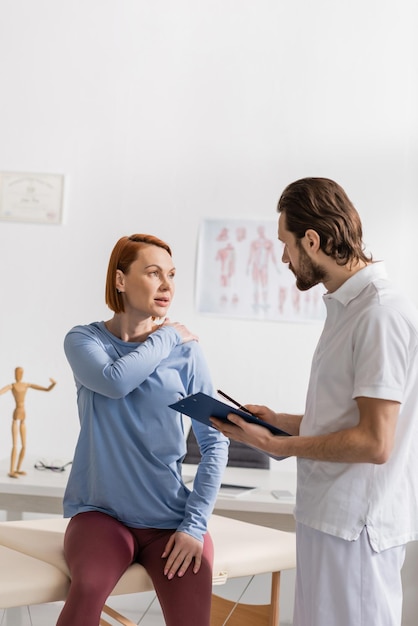 woman touching painful shoulder during consultation with physiotherapist holding clipboard