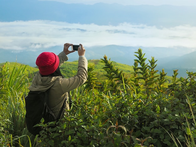 赤い帽子の女性観光バックパッカーが山でスマートフォンで写真を撮る