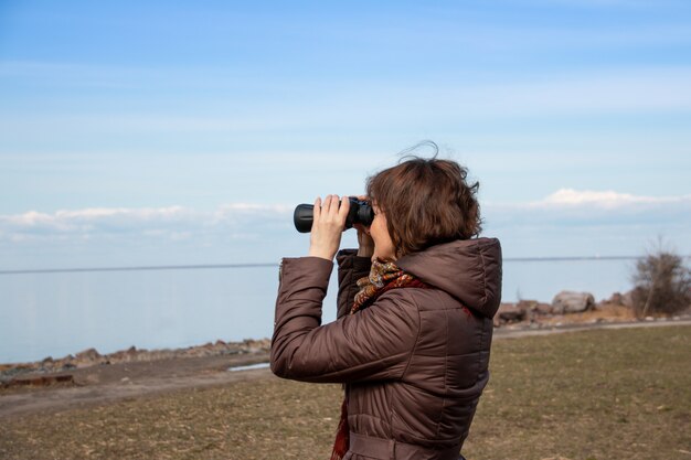 遠くの海で双眼鏡でみる、風景を楽しむ女性観光客。秋の時間。地平線、青い空を見て茶色のコートを着た孤独な女性。コピースペース