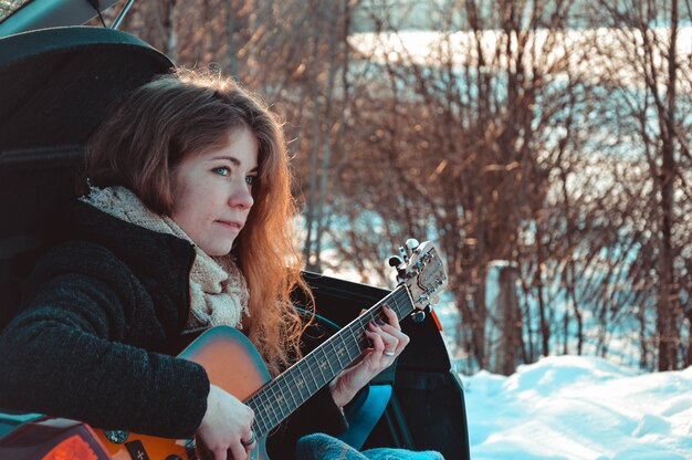 Turista della donna che si siede sulla macchina e suonare la chitarra è foresta invernale.