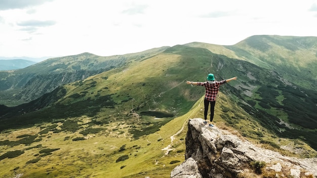 Woman tourist standing on edge of a cliff against background of green mountains and a lake