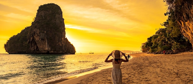 Woman tourist in white dress and hat walking on Phra nang Cave Beach at sunset Railay Krabi Thailand vacation travel summer Wanderlust and holiday concept