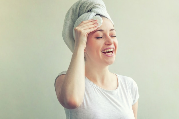 woman in towel on head removing make up with cotton pad on white background