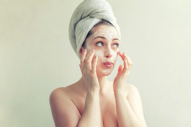 woman in towel on head with white nourishing mask or creme on face, white background isolated