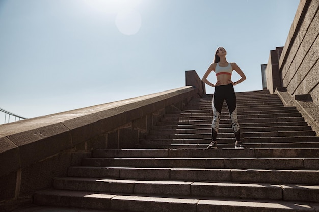 Woman in tracksuit with hands on waist stands on stone stairs on sunny summer day