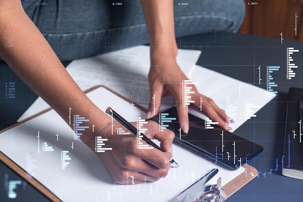 カジュアルウェアの女性トレーダーが、適切な投資のためにスマートフォンを使用して株式市場のトレンドを調査するためにいくつかの見積もりを書き留めていますウェルスマネジメントのコンセプトホログラム外国為替チャートのクローズアップショット