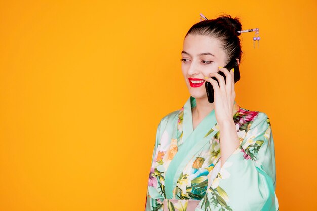 Woman in traditional japanese kimono happy and positive smiling while talking on mobile phone on orange